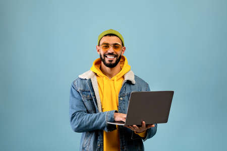 Successful arab freelancer guy using laptop computer and smiling to camera while standing over blue backgroundの写真素材