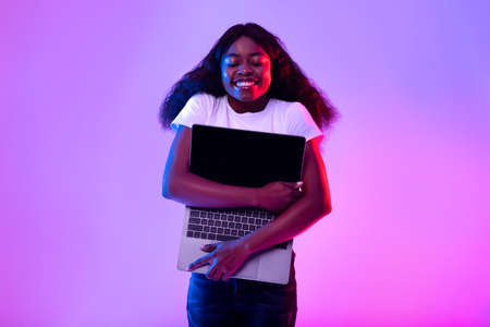Technology lover. Cherful black woman embracing laptop computer with empty screen in neon light, mockup for websiteの写真素材
