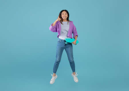 I like teenage culture. Joyful asian lady with skateboard jumping and showing thumb up over blue...