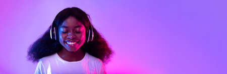 Pretty young black woman in headphones listening to music with closed eyes in neon light, panoramaの写真素材
