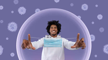 Happy black millennial guy showing protective mask after vaccination in bubble, protected from virusesの写真素材