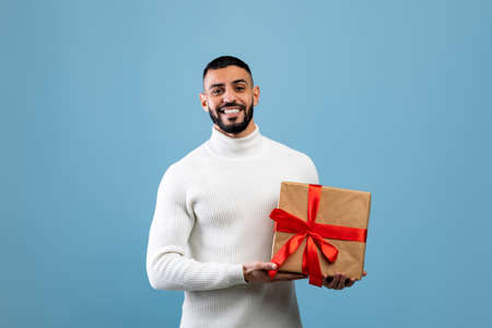 Best gift for you. Happy young arab man holding wrapped present box, smiling to camera, standing over blue backgroundの写真素材