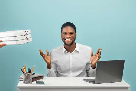 Excited black man working at desk with laptop, receiving food delivery, courier holding cardboard...