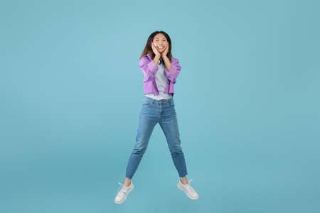 Shocking sale. Surprised amazed young asian lady jumping in air and touching face with amazement, blue backgroundの写真素材