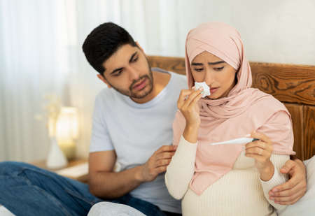 Illness during pregnancy. Loving muslim husband taking care about his sick pregnant wife, woman measuring temperatureの写真素材