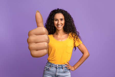 Cheerful young brunette woman gesturing thumb up over purple background