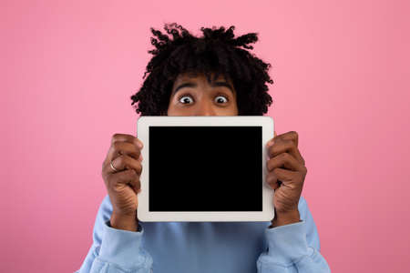 Shocked black teenager covering face with empty tablet computer, offering mockup for your app or website designの写真素材