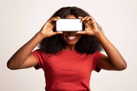 Mobile App. Cheerful Black Woman Covering Eyes With Blank Smartphone, Mockup