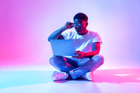 Shocked black guy sitting cross legged with laptop, touching his glasses, winning online lottery in neon lightの写真素材