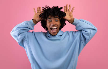 Cool African American teen guy making silly gesture with his hands, grimacing and having fun on pink studio backgroundの写真素材