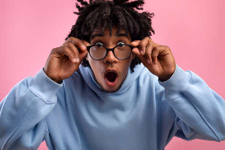 Shocked young black teen guy opening mouth in awe, touching eyeglasses, looking at camera on pink backgroundの写真素材