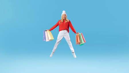 Excited young lady jumping with bags at blue studio background