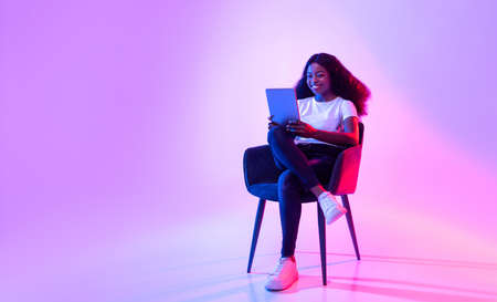 Cheerful black lady using tablet, studying or working remotely, sitting in armchair in neon light, copy spaceの写真素材