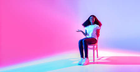 Happy black lady sitting on chair and offering empty space for your advertisement in neon light,...
