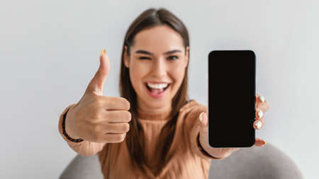 Young woman gesturing thumb up, showing cellphone with empty screen, winking and recommending new mobile app, mockupの写真素材