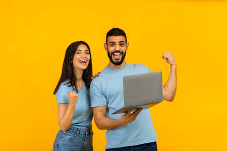 Overjoyed arab couple holding laptop and celebrating success with clenched fists, lucky winners with...
