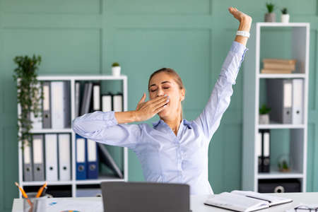Unproductive worker. Young lady yawning and stretching hands while sitting at workplace in modern...