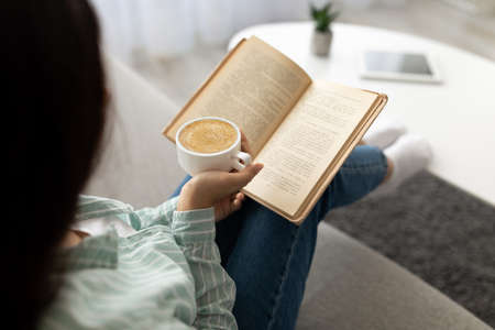 Unrecognizable Indian woman reading book, drinking fesh cappuccino at home. Hobbies and pastimes...