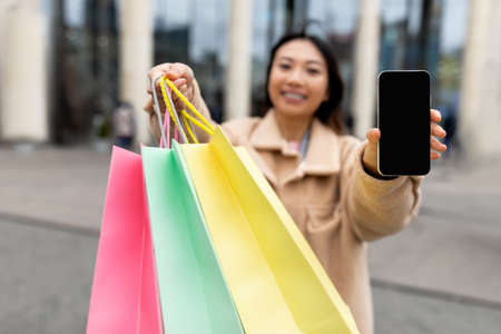 Happy young Asian woman showing shopper bags and smartphone with empty screen near big mall...