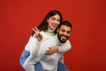 Joyful arab couple having fun and showing peace sign, happy guy piggybacking his wife and smiling, red backgroundの写真素材