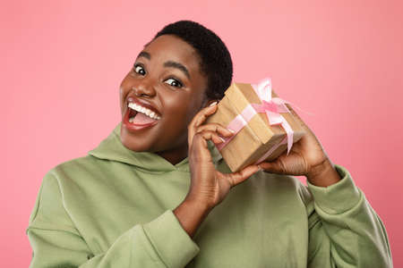 Excited Black Woman Holding Present Box Near Ear, Pink Backgroundの写真素材