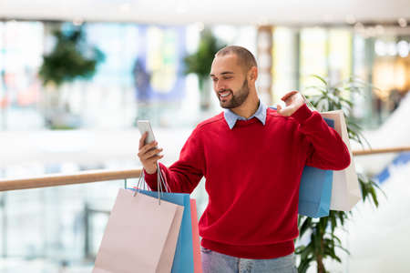 Positive young guy with gift bags using cellphone at mall, taking selfie or photo, having video call, free spaceの写真素材