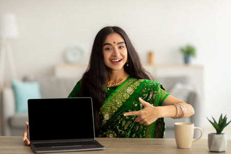 Cheerful Indian woman in green sari dress pointing at laptop computer with empty screen at home, mockup for websiteの写真素材