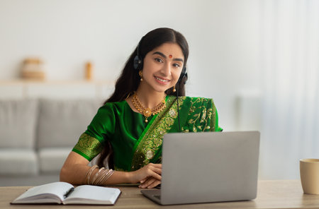 Indian woman in sari dress wearing headphones, having online meeting or web conference on laptop at homeの写真素材