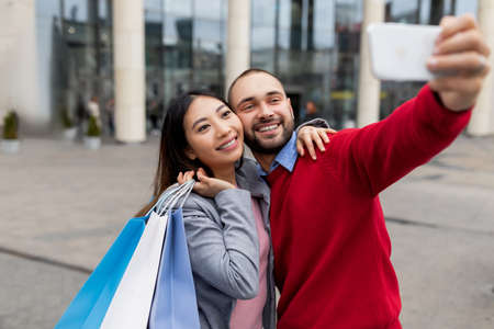 Loving young diverse couple with gift bags taking selfie near huge shopping centreの写真素材