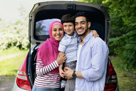 Happy Muslim Family With Little Son Posing Near Car With Open Trunkの写真素材