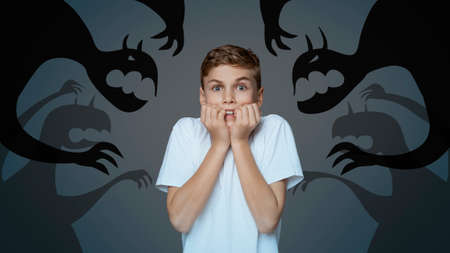 Afraid little boy biting nails in panic, scared of monster shadows on dark background, collage....