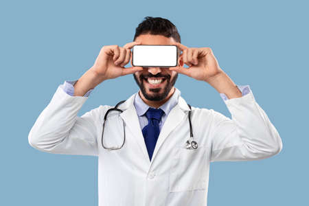 Male Doctor Holding Smartphone In Front Of Eyes, Blue Backgroundの写真素材