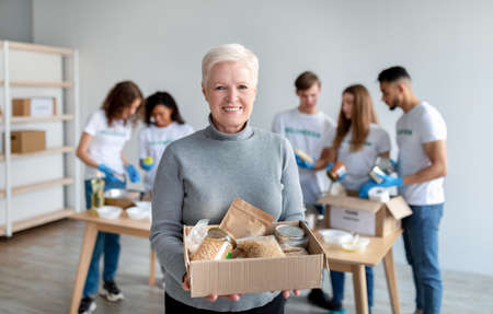 Happy senior woman holding box with donations food and smiling at camera while group of volunteers packing boxesの写真素材