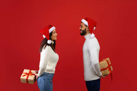 Happy loving arab spouses holding New Year presents behind their backs, exchanging gifts over red...