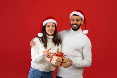 Merry Christmas. Cheerful arab spouses holding gift box, wearing Santa hats and smiling to camera over red backgroundの写真素材
