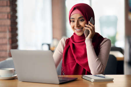 Beautiful muslim woman working online, having phone conversation, using laptopの写真素材