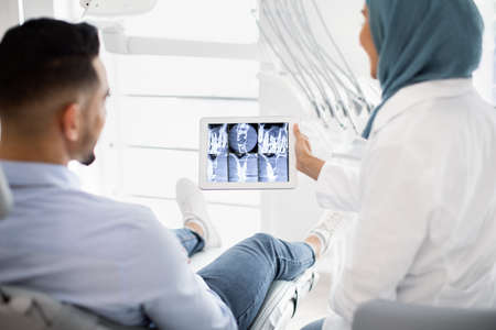 Muslim Female Dentist Showing Teeth X-ray On Digital Tablet To Male Patient
