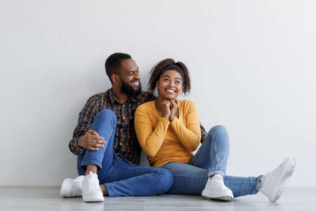 Cheerful young black husband and wife sitting on floor in empty room and enjoying moving to new homeの写真素材