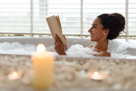 Lovely young woman lying in foamy bath, reading book in relaxing atmosphere with candles, indoors. Copy spaceの写真素材