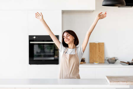 Relaxed young lady raising hands and feeling excited cooking alone in modern kitchen interior, having funの写真素材
