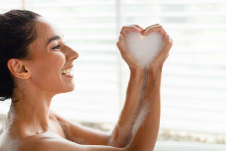 Happy young female sitting in bubble bath, making foamy heart with her hands at homeの写真素材