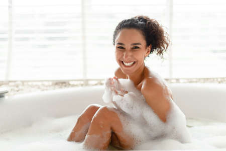 Portrait of smiling millennial woman covered in foam sitting in hot bubble bath at home, copy spaceの写真素材