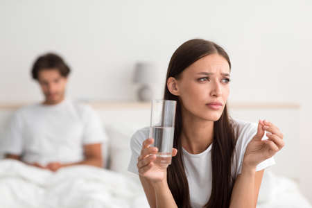 Sad young european lady hold pill and glass of water, husband waiting for wife in bed in bedroomの写真素材