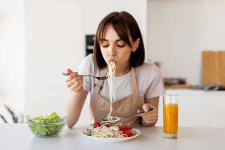 Young housewife tasting spaghetti and vegetable salad, enjoying delicious lunch at home, sitting in modern kitchenの写真素材