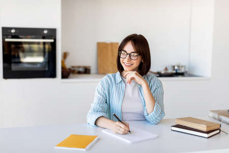 Keep diary, write down thoughts and ideas. Dreamy young lady sitting in kitchen and writing in...