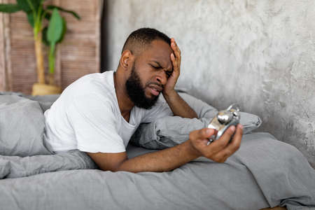 Black man suffering from insomnia looking at clock in bedの写真素材
