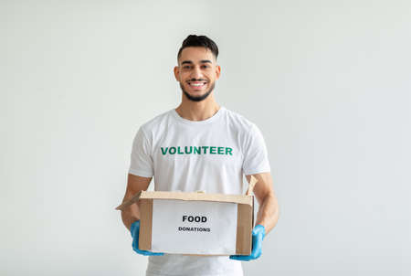 Portrait of young arab man wearing volunteer t-shirt, gloves, holding donations box, standing on light backgroundの写真素材