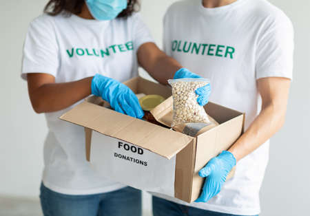 Female and male volunteers in protective gloves packing beans in cardboard box, working together on donation projectの写真素材