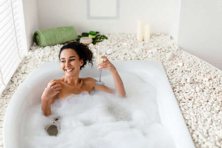 Happy young woman drinking champagne in bubble bath, celebrating holiday with alcoholic beverage in...