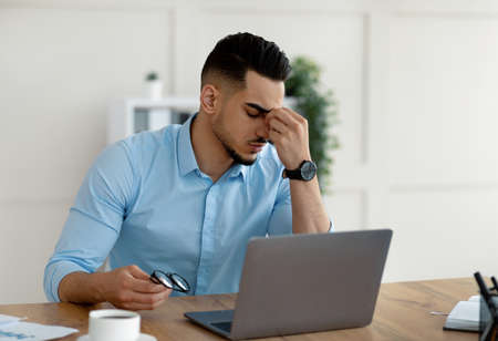 Millennial Arab entrepreneur having tired irritated eyes, overworking on laptop, exhausted from online job at officeの写真素材